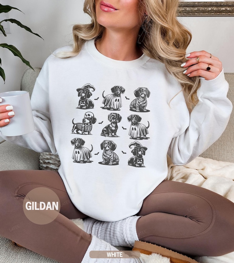 May include: White sweatshirt featuring a grid of dachshund illustrations in Halloween costumes. The dachshunds are depicted as a witch, ghost, skeleton, and more. The sweatshirt has a crew neck and long sleeves. The brand name "GILDAN" is visible.