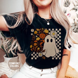 May include: Black t-shirt with a ghost graphic wearing a daisy crown, set against a checkered background. Floral elements in yellow and orange surround the ghost. The words "Comfort Colors" are printed on the shirt.