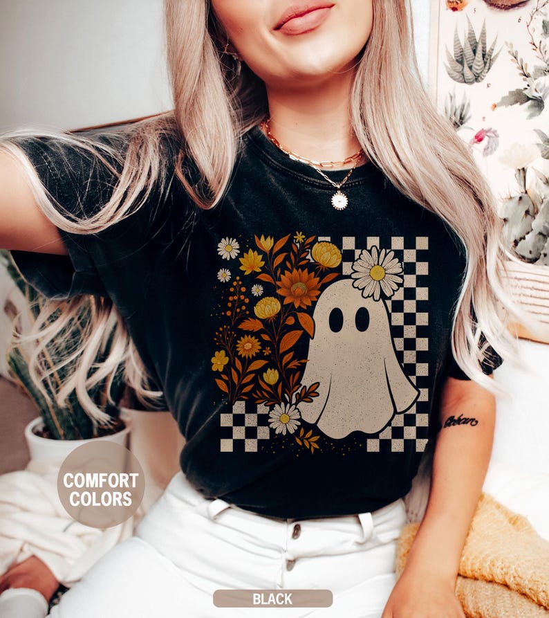May include: Black t-shirt with a ghost graphic wearing a daisy crown, set against a checkered background. Floral elements in yellow and orange surround the ghost. The words "Comfort Colors" are printed on the shirt.