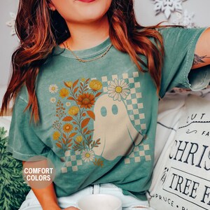 May include: Light green t-shirt featuring a ghost graphic with floral accents and a checkered background. The shirt has the words "Comfort Colors" and "Light Green" printed on it. The shirt is a comfortable, casual style.