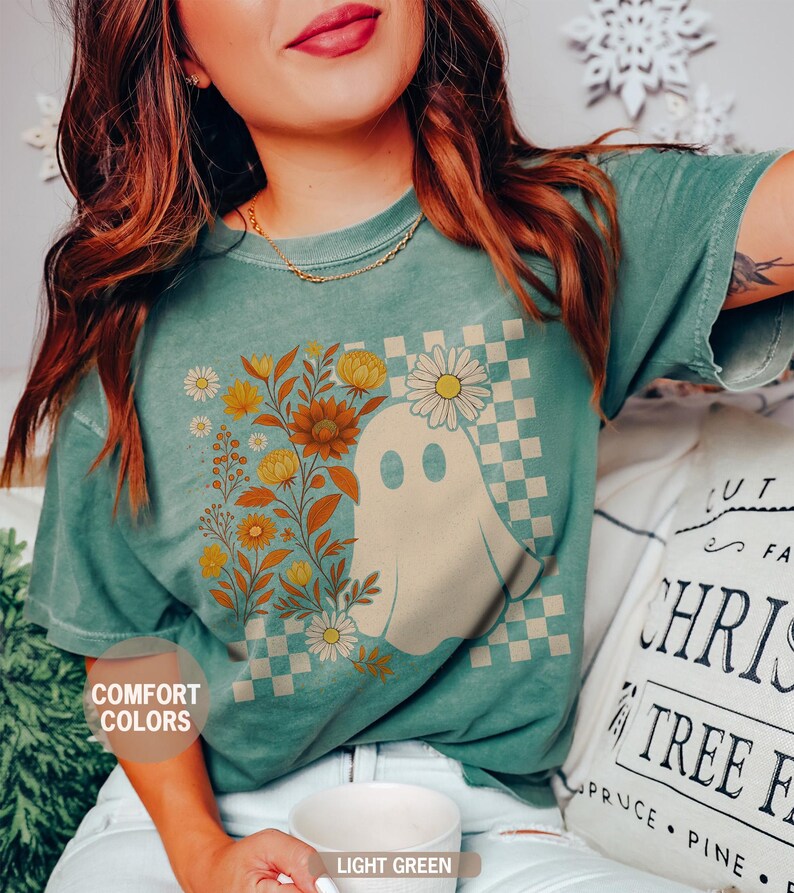 May include: Light green t-shirt featuring a ghost graphic with floral accents and a checkered background. The shirt has the words "Comfort Colors" and "Light Green" printed on it. The shirt is a comfortable, casual style.