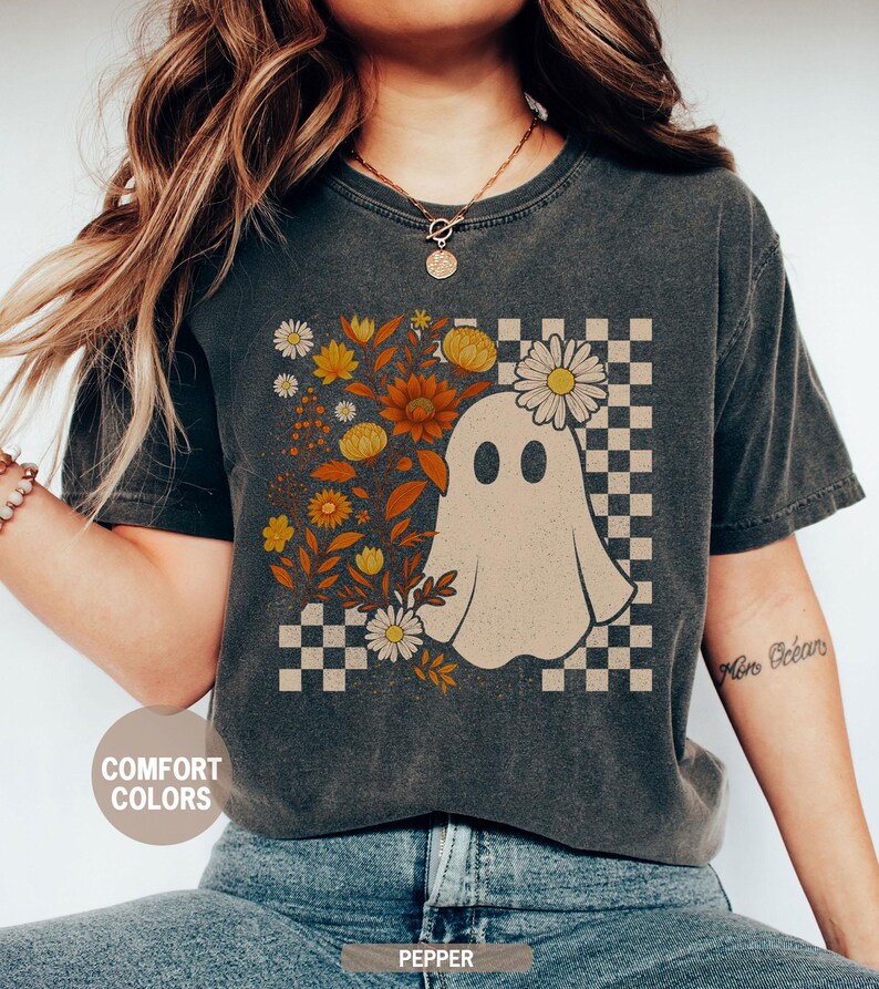 May include: Dark grey t-shirt featuring a ghost illustration with a floral design and a checkered background. The shirt has the words "Comfort Colors" and "Pepper" printed on it.