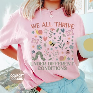 K&ouml;nnte beinhalten: Hellrosa T-Shirt mit dem Text "WE ALL THRIVE UNDER DIFFERENT CONDITIONS" und einem farbenfrohen Blumen- und Insektenmuster. Das Design umfasst Schmetterlinge, Bienen, Schnecken und eine Raupe. Das Shirt ist mit "COMFORT COLORS" und "BLOSSOM" gekennzeichnet.