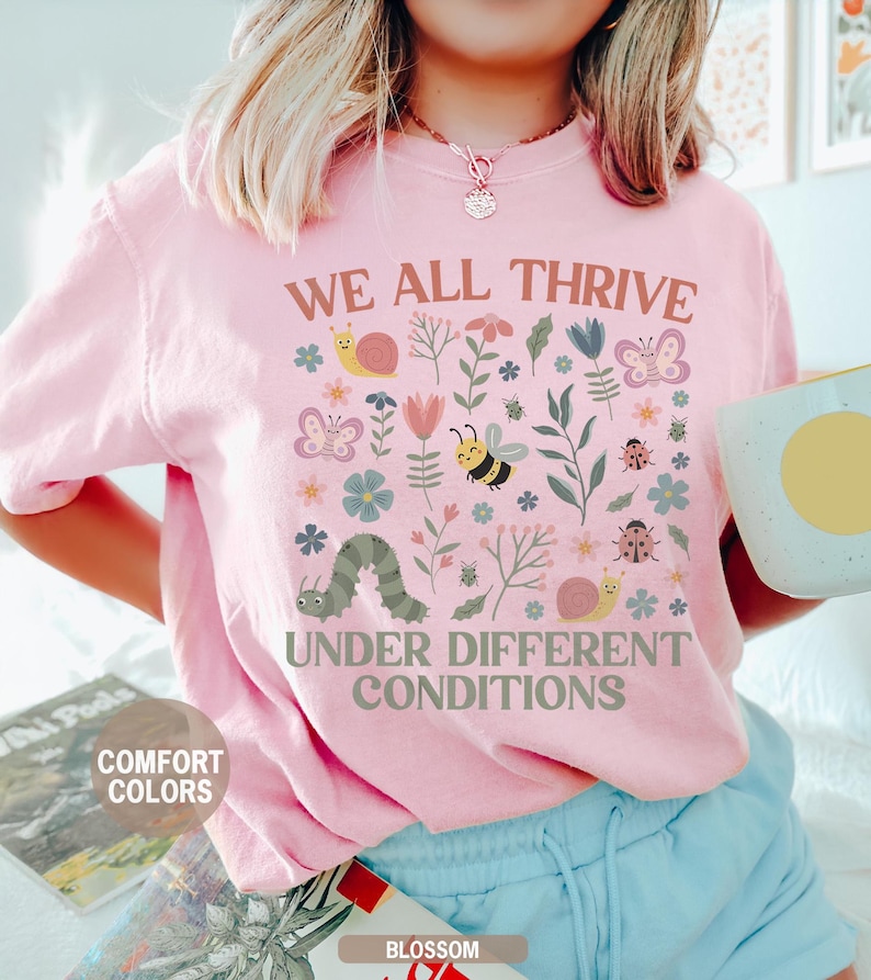 K&ouml;nnte beinhalten: Hellrosa T-Shirt mit dem Text "WE ALL THRIVE UNDER DIFFERENT CONDITIONS" und einem farbenfrohen Blumen- und Insektenmuster. Das Design umfasst Schmetterlinge, Bienen, Schnecken und eine Raupe. Das Shirt ist mit "COMFORT COLORS" und "BLOSSOM" gekennzeichnet.
