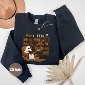 May include: Black sweatshirt with a Halloween-themed graphic. The graphic features a ghost reading in a chair in front of a bookshelf, with a black cat, pumpkins, and candles. The sweatshirt has a crew neck and long sleeves. The brand name "GILDAN" is visible.