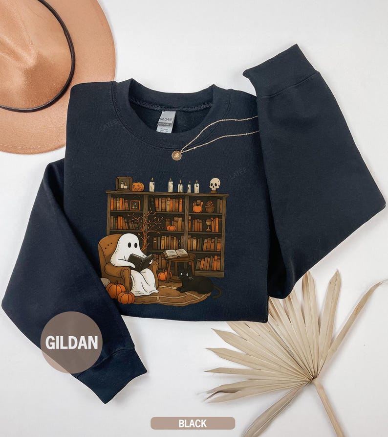 May include: Black sweatshirt with a Halloween-themed graphic. The graphic features a ghost reading in a chair in front of a bookshelf, with a black cat, pumpkins, and candles. The sweatshirt has a crew neck and long sleeves. The brand name "GILDAN" is visible.