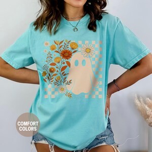 May include: A turquoise t-shirt featuring a floral ghost design. The graphic includes a beige ghost with daisy eyes, orange and yellow flowers, and a checkered background. The shirt is labeled "Comfort Colors" and "Chalky Mint".