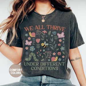 K&ouml;nnte beinhalten: Dunkelgraues T-Shirt mit dem Text "WE ALL THRIVE UNDER DIFFERENT CONDITIONS". Das Shirt zeigt ein farbenfrohes Blumendesign mit Schmetterlingen, Bienen, Schnecken und einer Raupe. Das Shirt ist mit "Comfort Colors" und "Pepper" gekennzeichnet.