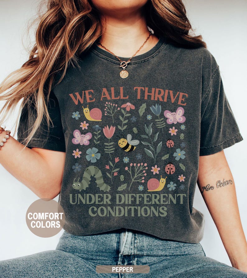 K&ouml;nnte beinhalten: Dunkelgraues T-Shirt mit dem Text "WE ALL THRIVE UNDER DIFFERENT CONDITIONS". Das Shirt zeigt ein farbenfrohes Blumendesign mit Schmetterlingen, Bienen, Schnecken und einer Raupe. Das Shirt ist mit "Comfort Colors" und "Pepper" gekennzeichnet.