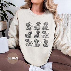 May include: Sand-colored sweatshirt featuring a grid of nine dachshund illustrations in Halloween costumes. The dachshunds are dressed as a witch, ghost, mummy, and skeleton. The brand name "GILDAN" is printed on the bottom left.