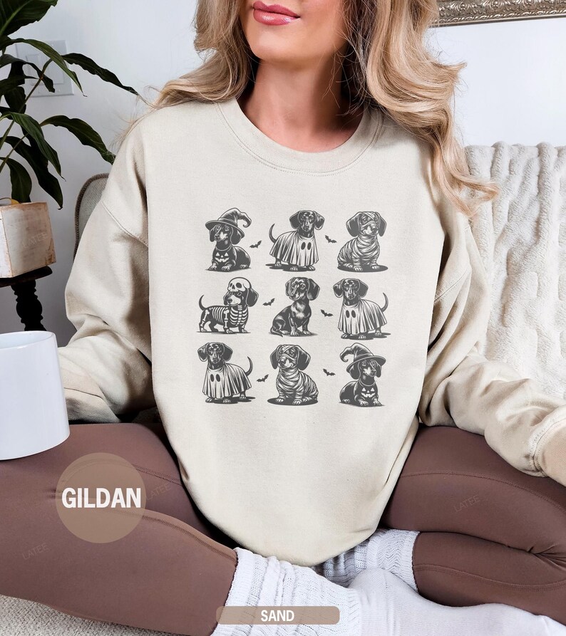 May include: Sand-colored sweatshirt featuring a grid of nine dachshund illustrations in Halloween costumes. The dachshunds are dressed as a witch, ghost, mummy, and skeleton. The brand name "GILDAN" is printed on the bottom left.