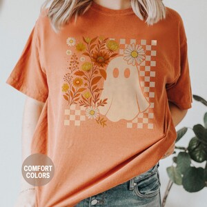 May include: Orange Comfort Colors t-shirt featuring a ghost design with flowers and a checkered background. The ghost is white, and the flowers are yellow and orange. The shirt has short sleeves and a round neckline.