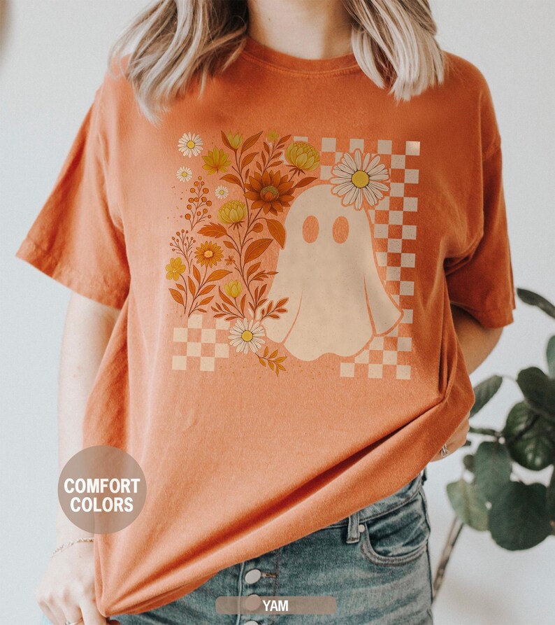 May include: Orange Comfort Colors t-shirt featuring a ghost design with flowers and a checkered background. The ghost is white, and the flowers are yellow and orange. The shirt has short sleeves and a round neckline.