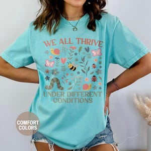 K&ouml;nnte beinhalten: Ein hellblaues T-Shirt mit den Worten "WE ALL THRIVE UNDER DIFFERENT CONDITIONS" in Pfirsich. Das Shirt zeigt eine farbenfrohe Grafik von Blumen, Insekten und einer Raupe. Das Shirt ist von der Marke Comfort Colors.
