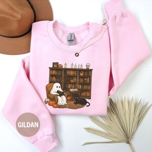 May include: Light pink Gildan sweatshirt featuring a Halloween-themed graphic. The graphic depicts a ghost reading in a cozy armchair in front of a bookshelf, with pumpkins, a black cat, and skulls. A gold necklace is draped on the sweatshirt.