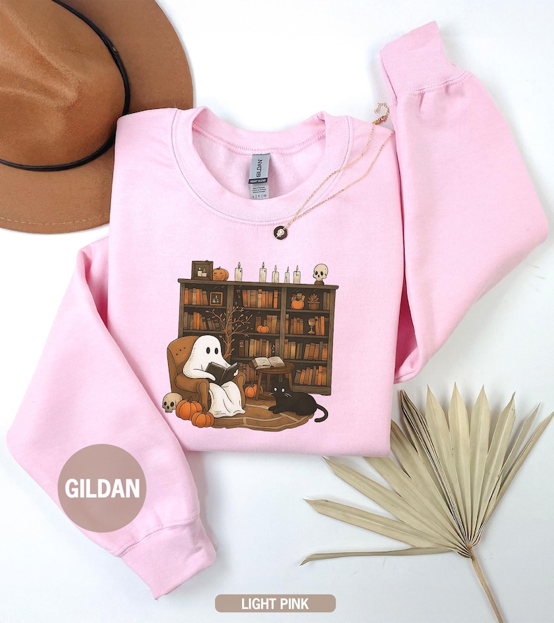 May include: Light pink Gildan sweatshirt featuring a Halloween-themed graphic. The graphic depicts a ghost reading in a cozy armchair in front of a bookshelf, with pumpkins, a black cat, and skulls. A gold necklace is draped on the sweatshirt.