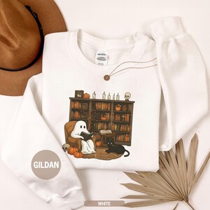 May include: White sweatshirt with a Halloween-themed graphic. The design features a ghost reading a book in a cozy armchair, a black cat, pumpkins, and a bookshelf. The word "WHITE" is printed at the bottom. The brand name "GILDAN" is printed on the sleeve.