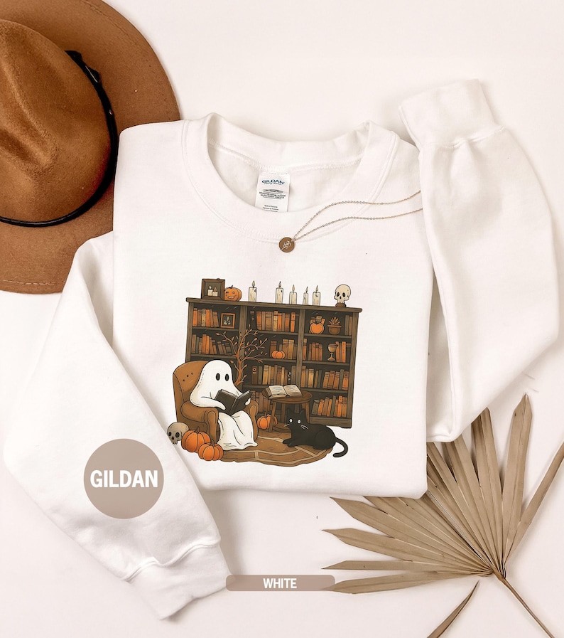 May include: White sweatshirt with a Halloween-themed graphic. The design features a ghost reading a book in a cozy armchair, a black cat, pumpkins, and a bookshelf. The word "WHITE" is printed at the bottom. The brand name "GILDAN" is printed on the sleeve.