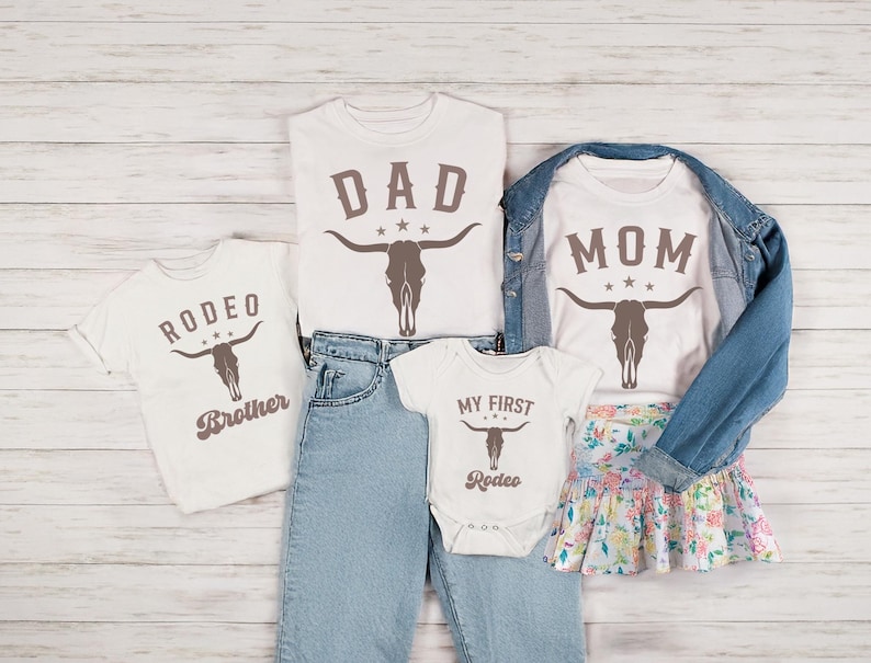 May include: Four white t-shirts with brown skull and crossbones designs. The shirts have the text 'DAD', 'MOM', 'RODEO BROTHER', and 'MY FIRST RODEO'. The shirts are displayed on a white wood background.