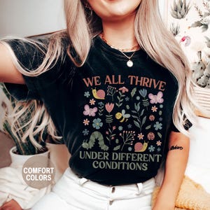 K&ouml;nnte beinhalten: Schwarzes T-Shirt mit dem Text "WE ALL THRIVE UNDER DIFFERENT CONDITIONS" und einem farbenfrohen Blumen- und Insektenmuster. Das Design umfasst Schmetterlinge, Bienen, Schnecken und eine Raupe. Das Shirt ist mit "COMFORT COLORS" und "BLACK" gekennzeichnet.