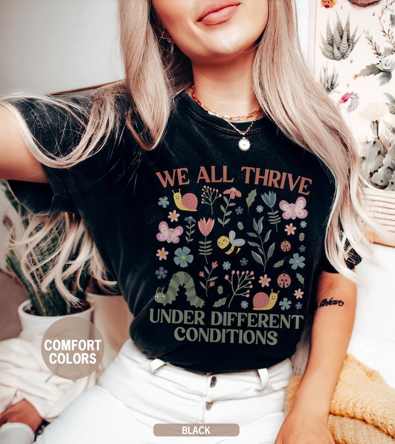 K&ouml;nnte beinhalten: Schwarzes T-Shirt mit dem Text "WE ALL THRIVE UNDER DIFFERENT CONDITIONS" und einem farbenfrohen Blumen- und Insektenmuster. Das Design umfasst Schmetterlinge, Bienen, Schnecken und eine Raupe. Das Shirt ist mit "COMFORT COLORS" und "BLACK" gekennzeichnet.