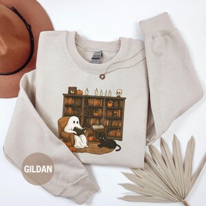 May include: Sand-colored sweatshirt featuring a graphic of a ghost reading in a cozy library setting with a black cat. The design includes pumpkins, candles, and a bookshelf. The sweatshirt has a crew neck and long sleeves.