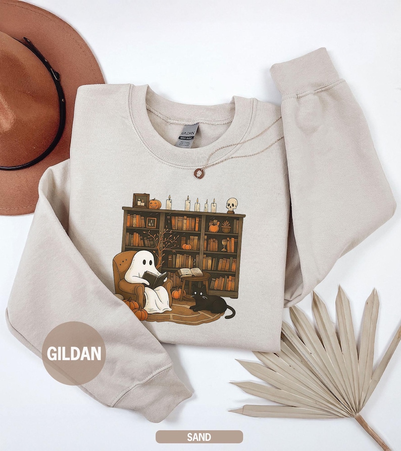 May include: Sand-colored sweatshirt featuring a graphic of a ghost reading in a cozy library setting with a black cat. The design includes pumpkins, candles, and a bookshelf. The sweatshirt has a crew neck and long sleeves.
