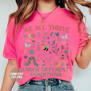 K&ouml;nnte beinhalten: Ein leuchtend pinkfarbenes T-Shirt mit dem Aufdruck "WE ALL THRIVE UNDER DIFFERENT CONDITIONS". Das Design umfasst Insekten, Blumen und eine Raupe. Das Shirt ist von der Marke Comfort Colors.