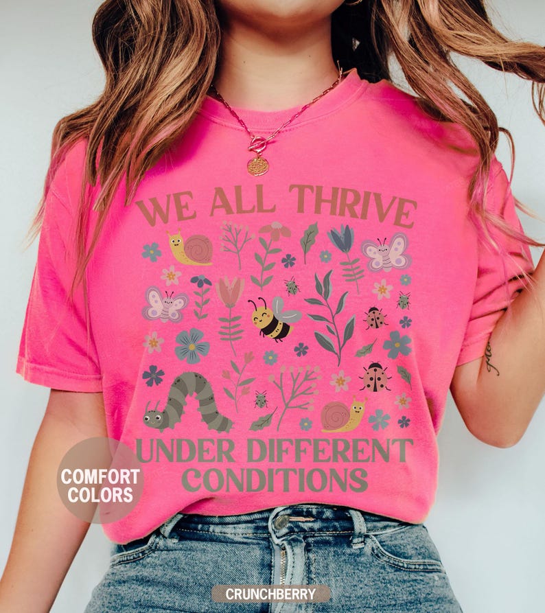 K&ouml;nnte beinhalten: Ein leuchtend pinkfarbenes T-Shirt mit dem Aufdruck "WE ALL THRIVE UNDER DIFFERENT CONDITIONS". Das Design umfasst Insekten, Blumen und eine Raupe. Das Shirt ist von der Marke Comfort Colors.