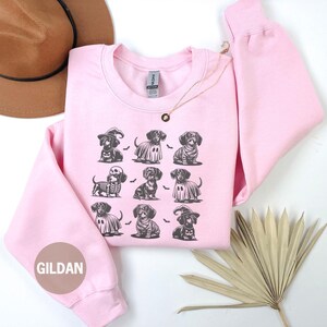 May include: Light pink sweatshirt featuring a repeating pattern of dachshunds in Halloween costumes. The dachshunds are dressed as a witch, ghost, skeleton, and more. The brand name "GILDAN" is printed on the sleeve. The words "LIGHT PINK" are printed at the bottom.