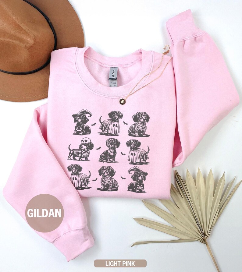 May include: Light pink sweatshirt featuring a repeating pattern of dachshunds in Halloween costumes. The dachshunds are dressed as a witch, ghost, skeleton, and more. The brand name "GILDAN" is printed on the sleeve. The words "LIGHT PINK" are printed at the bottom.