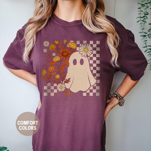 May include: Berry-colored t-shirt featuring a whimsical ghost design. The ghost is surrounded by floral elements and a checkered pattern. The shirt has the words "Comfort Colors" and "Berry" printed on it.