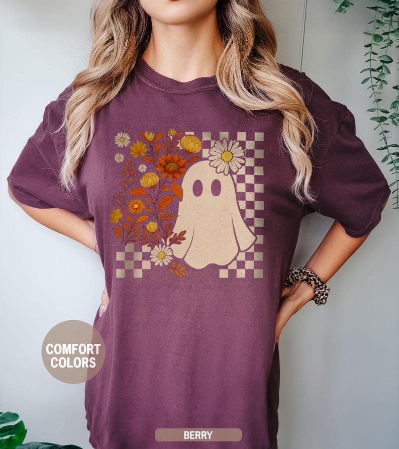 May include: Berry-colored t-shirt featuring a whimsical ghost design. The ghost is surrounded by floral elements and a checkered pattern. The shirt has the words "Comfort Colors" and "Berry" printed on it.