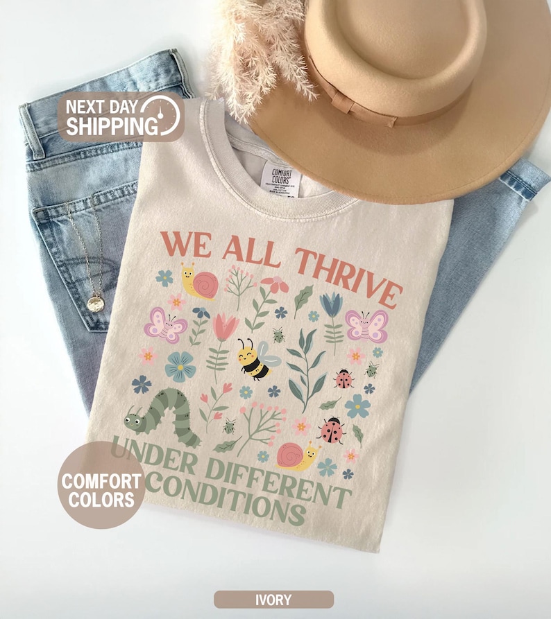 K&ouml;nnte beinhalten: Elfenbeinfarbenes T-Shirt mit dem Text "WE ALL THRIVE UNDER DIFFERENT CONDITIONS" und farbenfrohen Illustrationen von Insekten und Blumen. Das Shirt liegt auf einer blauen Jeans und einem beigen Hut.
