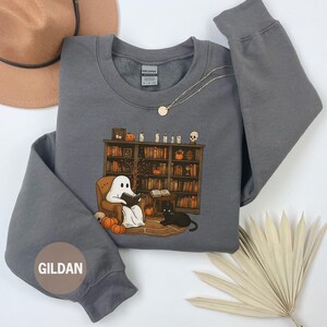 May include: Dark heather gray sweatshirt with a graphic of a ghost reading in a cozy library setting, complete with pumpkins, a black cat, and a bookshelf. The sweatshirt has the brand name "GILDAN" on the sleeve.