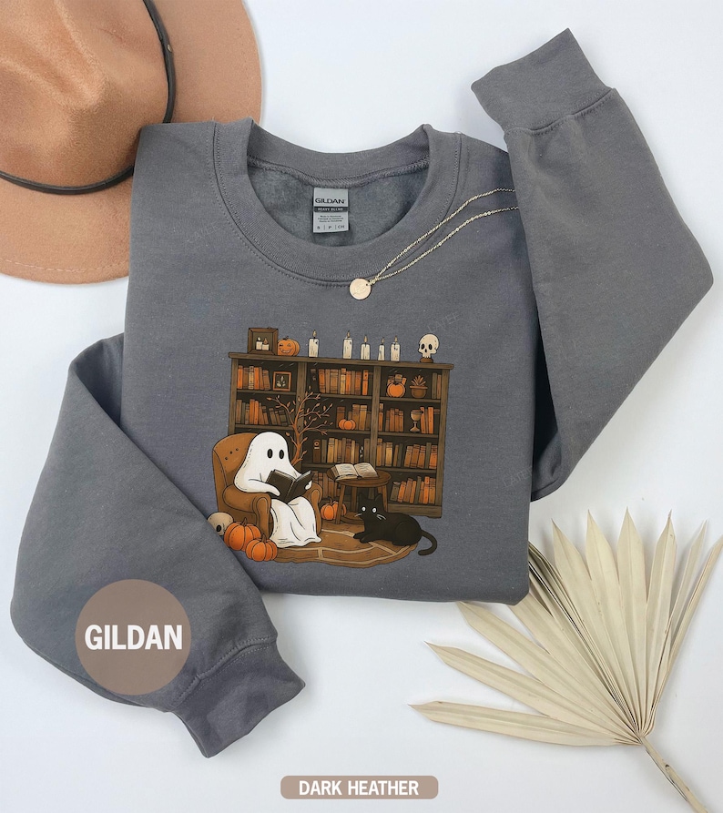 May include: Dark heather gray sweatshirt with a graphic of a ghost reading in a cozy library setting, complete with pumpkins, a black cat, and a bookshelf. The sweatshirt has the brand name "GILDAN" on the sleeve.