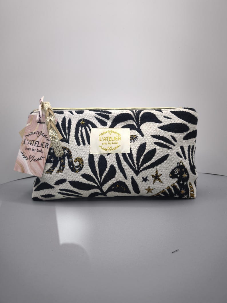 Pode incluir: Uma bolsa de cosm&eacute;ticos retangular com um padr&atilde;o de folhas em preto e branco. A bolsa tem um z&iacute;per dourado e uma etiqueta com o texto "L'ATELIER sous les toits". A bolsa &eacute; decorada com estrelas douradas e um animal preto e dourado.