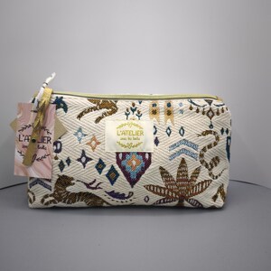 Pode incluir: Bolsa de cosm&eacute;ticos retangular creme com z&iacute;per dourado e padr&otilde;es bordados intrincados em marrom, azul e roxo. A bolsa apresenta uma etiqueta com texto e uma etiqueta decorativa.