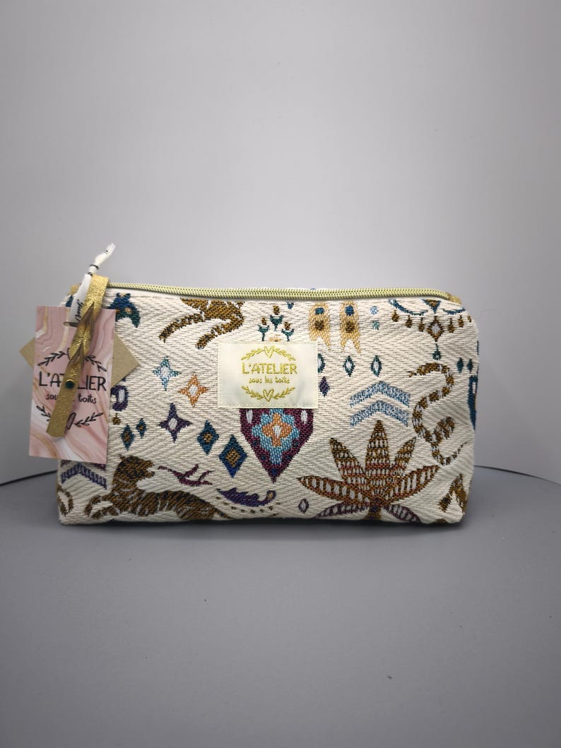 Pode incluir: Bolsa de cosm&eacute;ticos retangular creme com z&iacute;per dourado e padr&otilde;es bordados intrincados em marrom, azul e roxo. A bolsa apresenta uma etiqueta com texto e uma etiqueta decorativa.