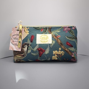 Pode incluir: Bolsa de cosm&eacute;ticos retangular azul-petr&oacute;leo com um padr&atilde;o floral e de p&aacute;ssaros. A bolsa apresenta um z&iacute;per dourado e uma etiqueta com o texto "L'ATELIER sous les toits". O design inclui flores vermelhas, amarelas e rosas e um p&aacute;ssaro castanho.