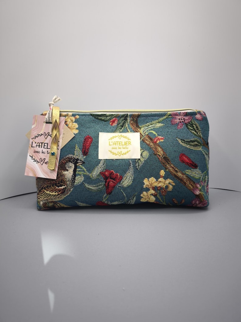 Pode incluir: Bolsa de cosm&eacute;ticos retangular azul-petr&oacute;leo com um padr&atilde;o floral e de p&aacute;ssaros. A bolsa apresenta um z&iacute;per dourado e uma etiqueta com o texto "L'ATELIER sous les toits". O design inclui flores vermelhas, amarelas e rosas e um p&aacute;ssaro castanho.