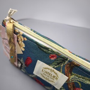 Pode incluir: Estojo turquesa com um padr&atilde;o floral com flores vermelhas e amarelas. Possui um z&iacute;per dourado e uma etiqueta com a inscri&ccedil;&atilde;o "L'ATELIER sous les toits". Uma fita dourada est&aacute; presa ao z&iacute;per.
