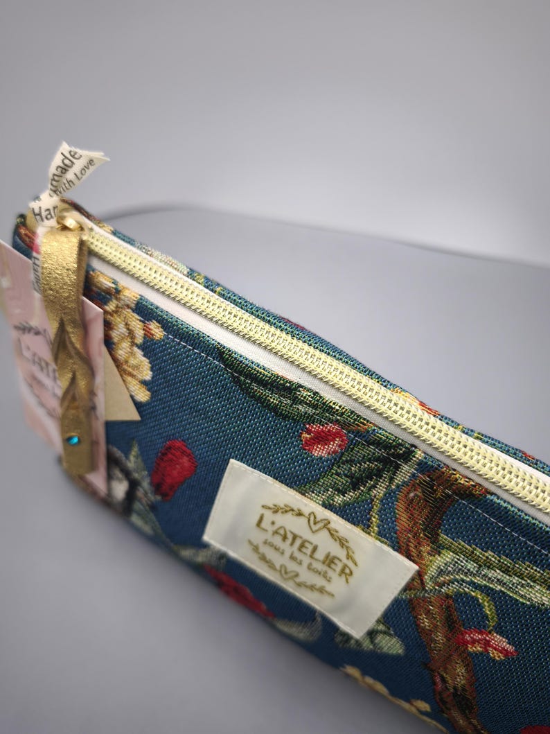 Pode incluir: Estojo turquesa com um padr&atilde;o floral com flores vermelhas e amarelas. Possui um z&iacute;per dourado e uma etiqueta com a inscri&ccedil;&atilde;o "L'ATELIER sous les toits". Uma fita dourada est&aacute; presa ao z&iacute;per.