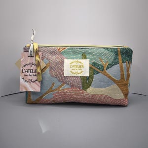 Pode incluir: Necessaire retangular de tape&ccedil;aria com um desenho de paisagem em tons de azul, verde, rosa e castanho. A bolsa tem um fecho de correr dourado e uma etiqueta que diz "L'ATELIER sous les toits".