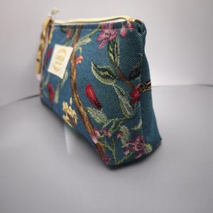 Pode incluir: Bolsa de cosm&eacute;ticos de tecido azul-petr&oacute;leo com um padr&atilde;o floral em rosa, vermelho e amarelo. A bolsa tem um z&iacute;per dourado e uma pequena etiqueta. A bolsa &eacute; retangular e tem um topo arredondado.