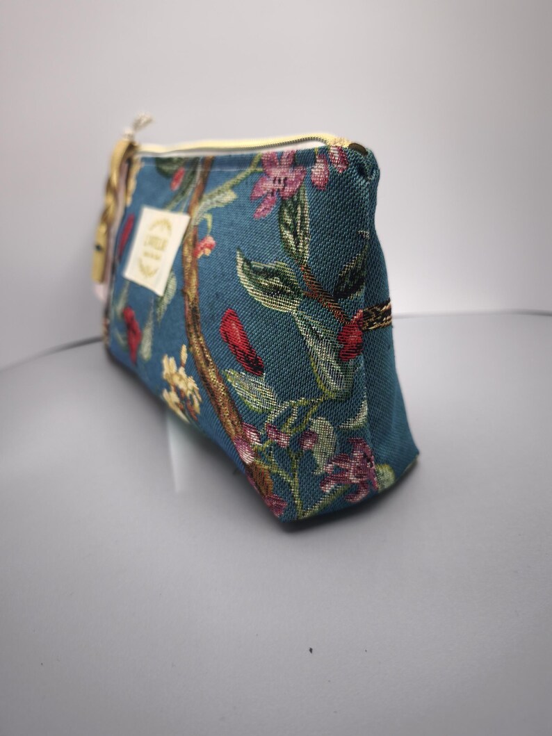 Pode incluir: Bolsa de cosm&eacute;ticos de tecido azul-petr&oacute;leo com um padr&atilde;o floral em rosa, vermelho e amarelo. A bolsa tem um z&iacute;per dourado e uma pequena etiqueta. A bolsa &eacute; retangular e tem um topo arredondado.