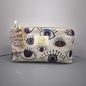Pode incluir: Bolsa retangular creme com um padr&atilde;o bordado de olhos em azul, vermelho e dourado. A bolsa tem um z&iacute;per dourado e uma etiqueta com o texto "L'ATELIER sous les toits".