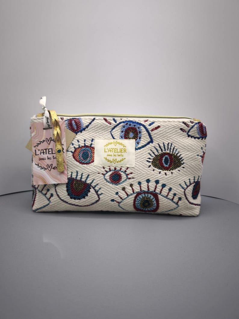 Pode incluir: Bolsa retangular creme com um padr&atilde;o bordado de olhos em azul, vermelho e dourado. A bolsa tem um z&iacute;per dourado e uma etiqueta com o texto "L'ATELIER sous les toits".