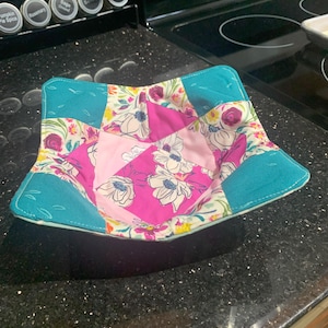 May include: A teal, pink and white floral fabric bowl cosy. The bowl cosy is made of patchwork fabric with a floral pattern.