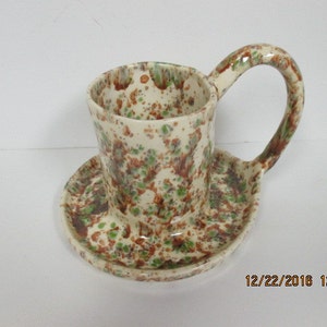 May include: A ceramic cup and saucer set with a speckled brown, green, and white glaze. The cup has a large handle and the saucer has a unique shape.